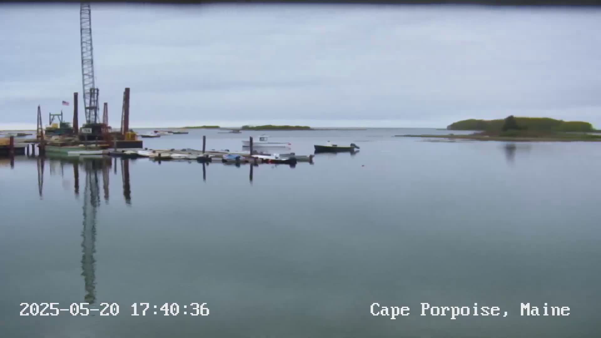Several boats are moored at a calm harbor under an overcast sky, with a construction crane visible in the background.