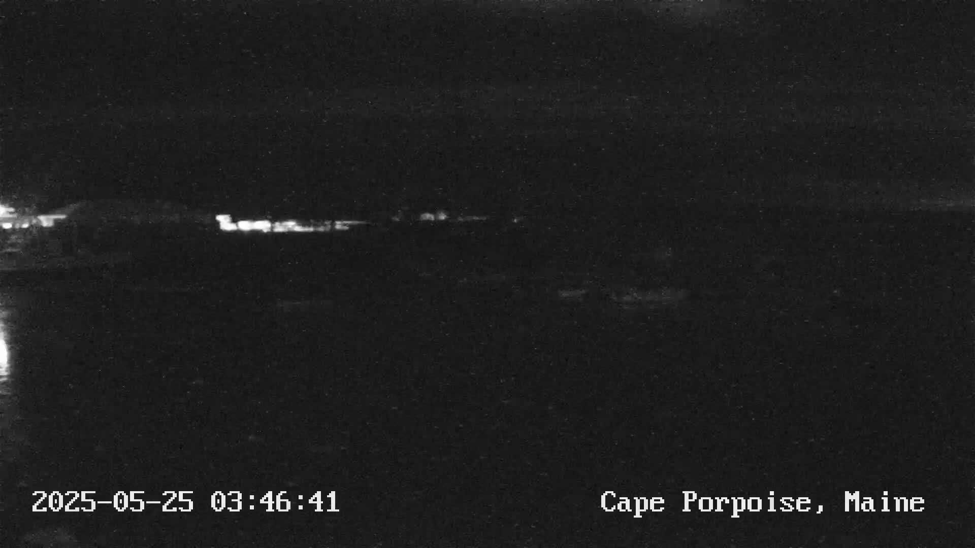 A low-light nighttime image shows a dark, mostly featureless landscape with a faint line of lights in the distance.