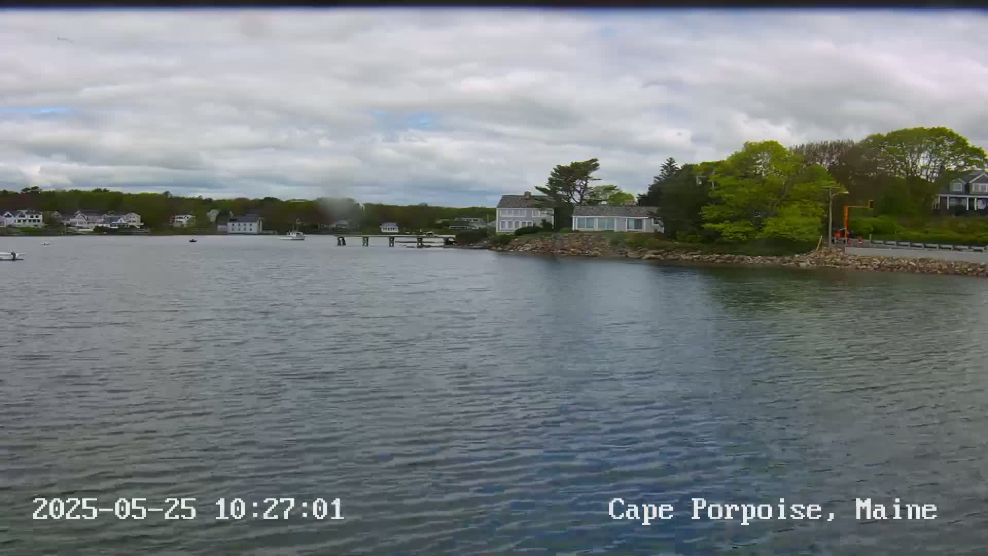 A calm body of water shows several houses along a shoreline under a cloudy sky.