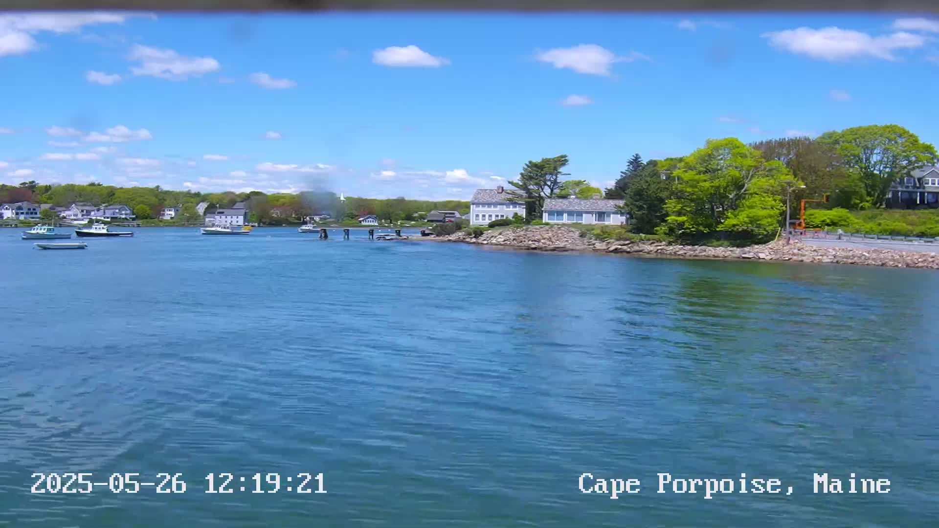 A calm body of water with several boats and houses on the shoreline under a partly cloudy blue sky.