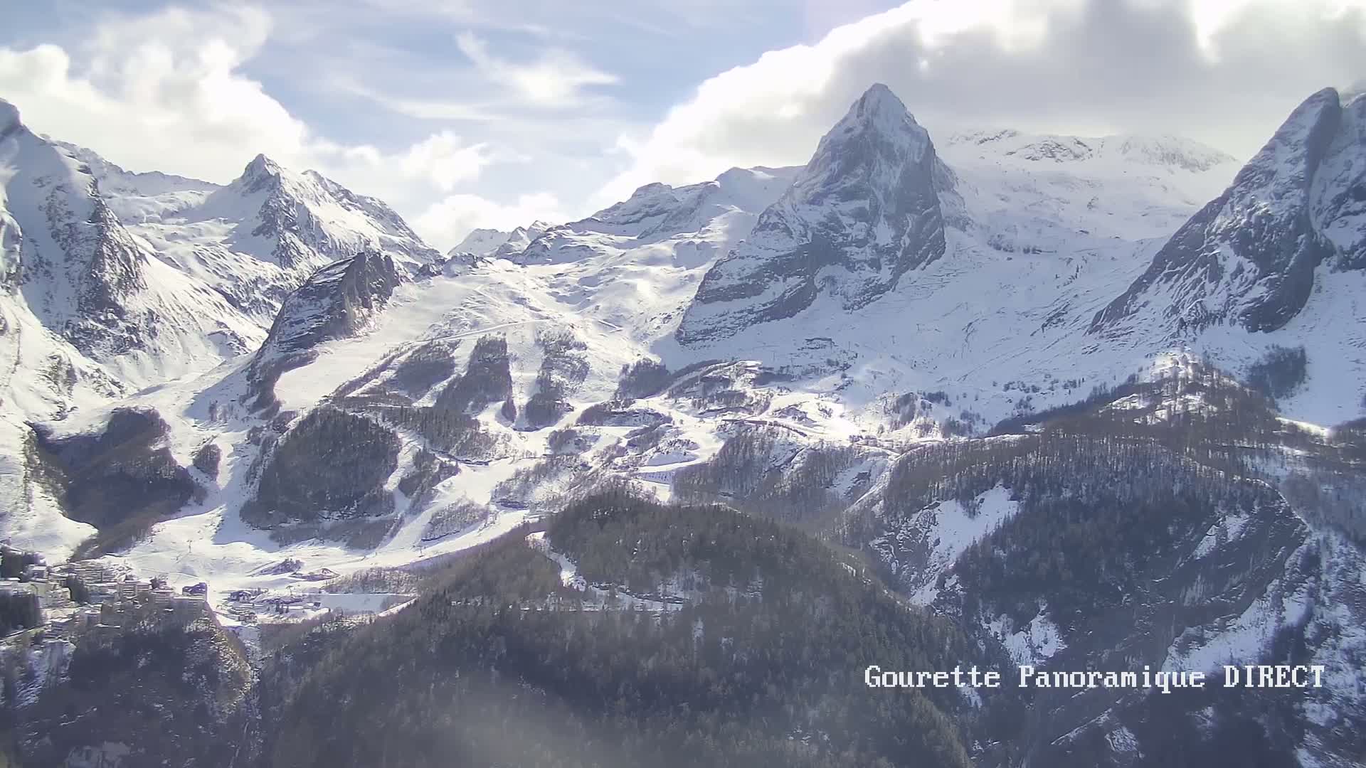 A snow-covered mountain range with a valley below, under a partly cloudy sky.