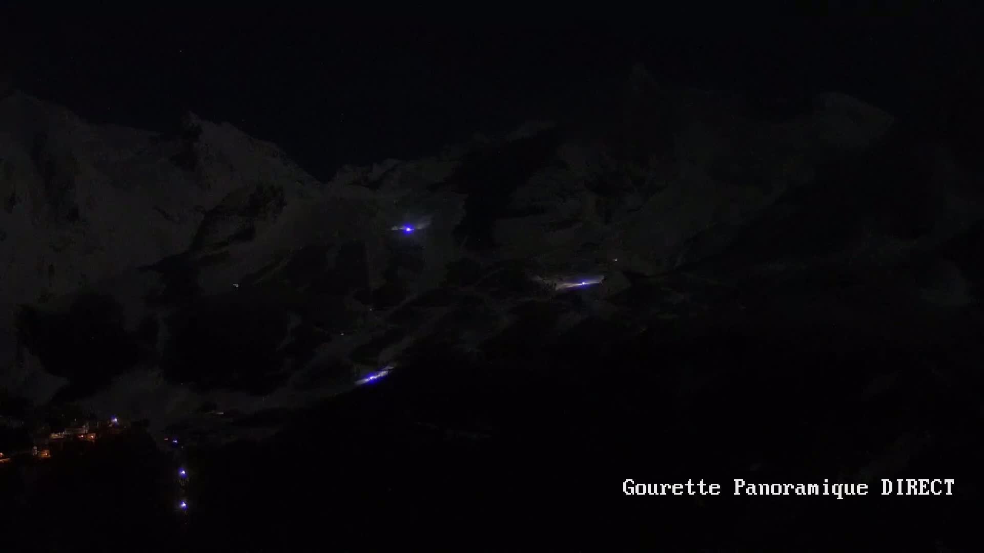 A nighttime view of snow-covered mountains with a few small lights visible below and on the mountainside.