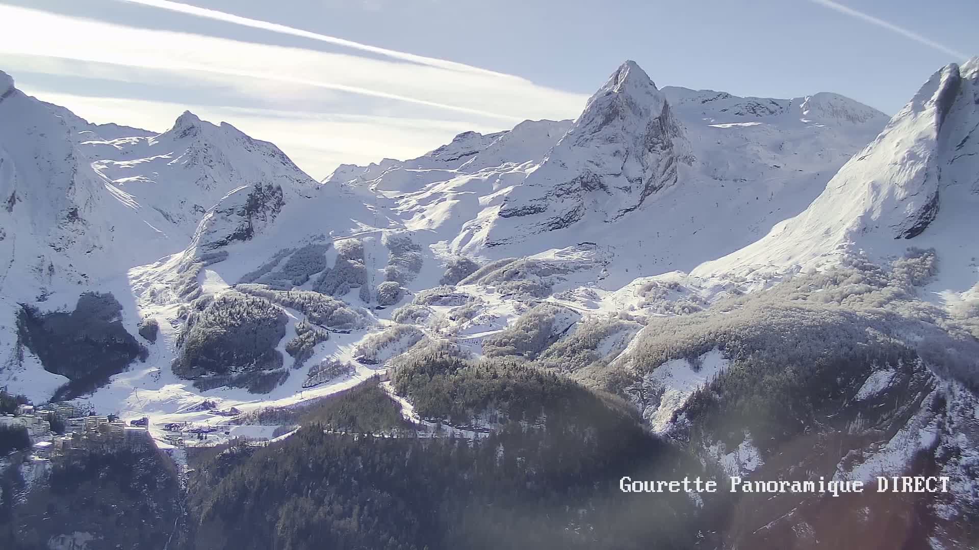 A snow-covered mountain range with a ski resort nestled in a valley below, under a bright, sunny sky with contrails.