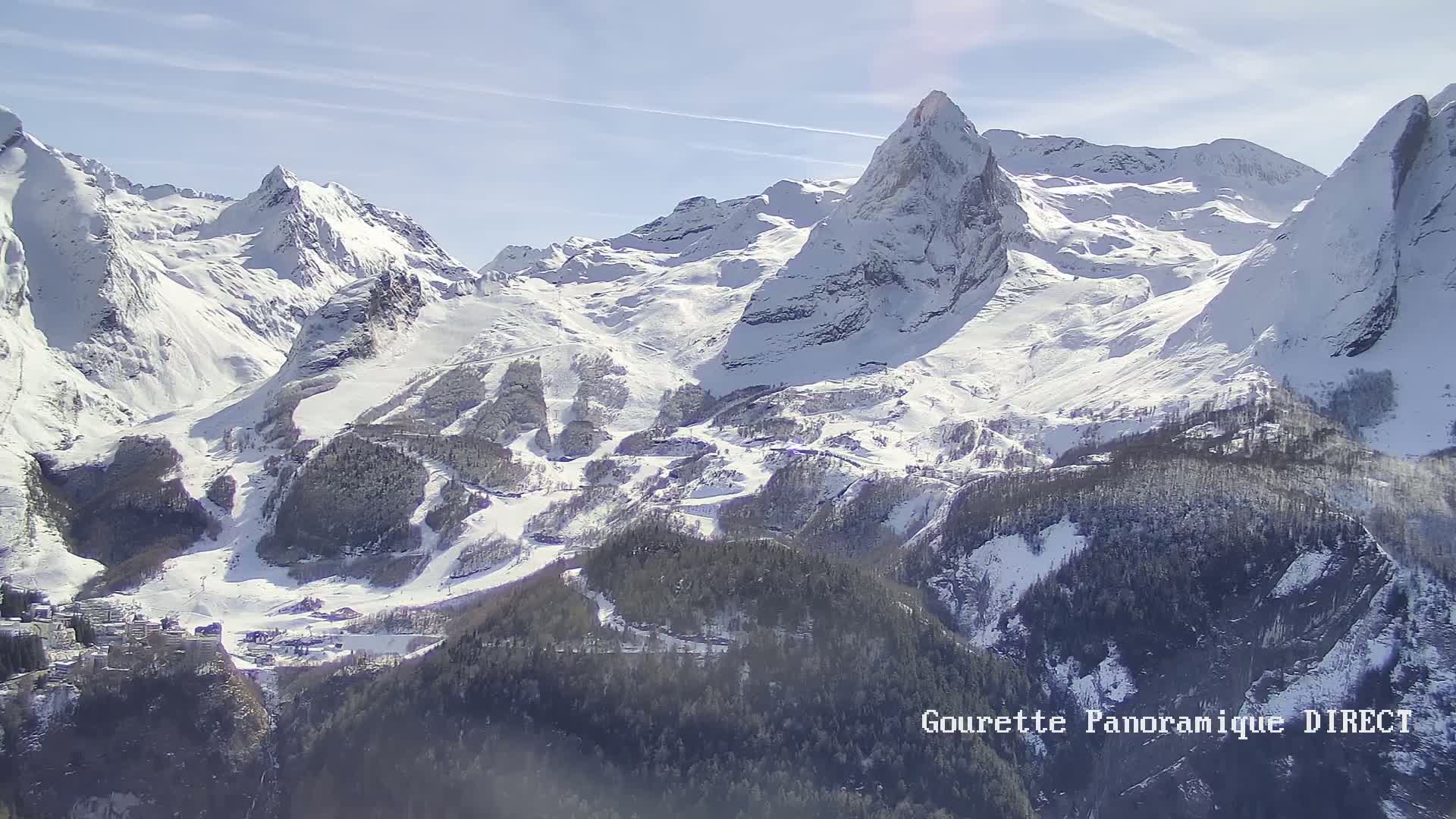 A snow-covered mountain range is seen on a sunny day, with a valley of evergreen trees in the foreground.