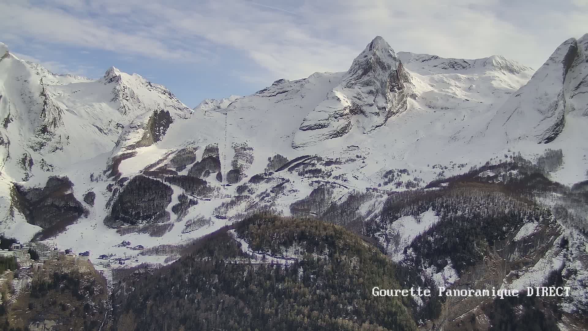 A snow-covered mountain range with a village nestled in the valley below under a partly cloudy sky.