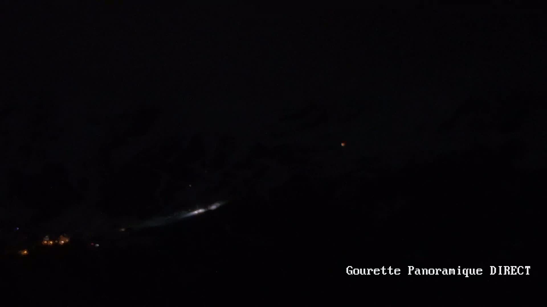 A nighttime long-exposure shot shows a bright streak of light across a dark sky, with a small, bright light and a few distant lights visible near the horizon.