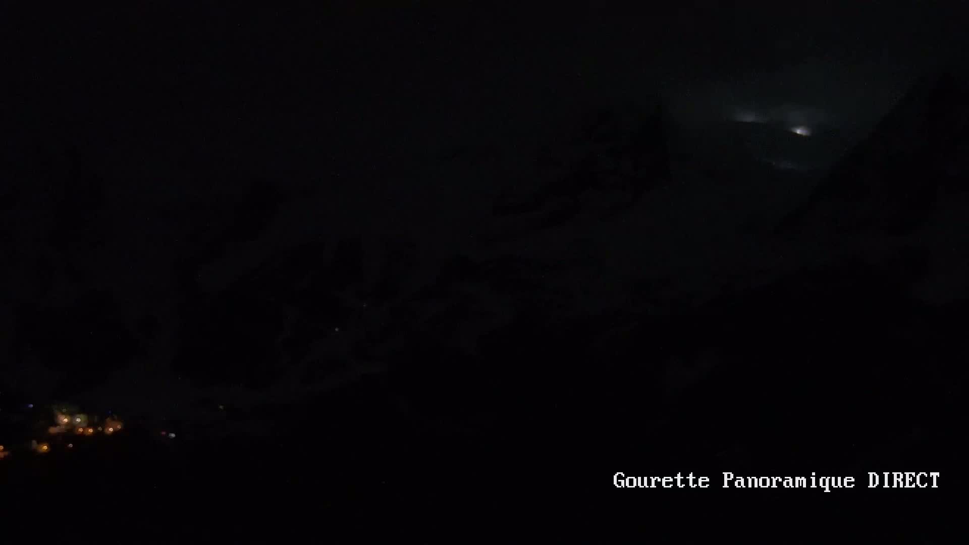 A nighttime view of a moon partially obscured by clouds above a dark landscape with faintly visible lights in the distance.