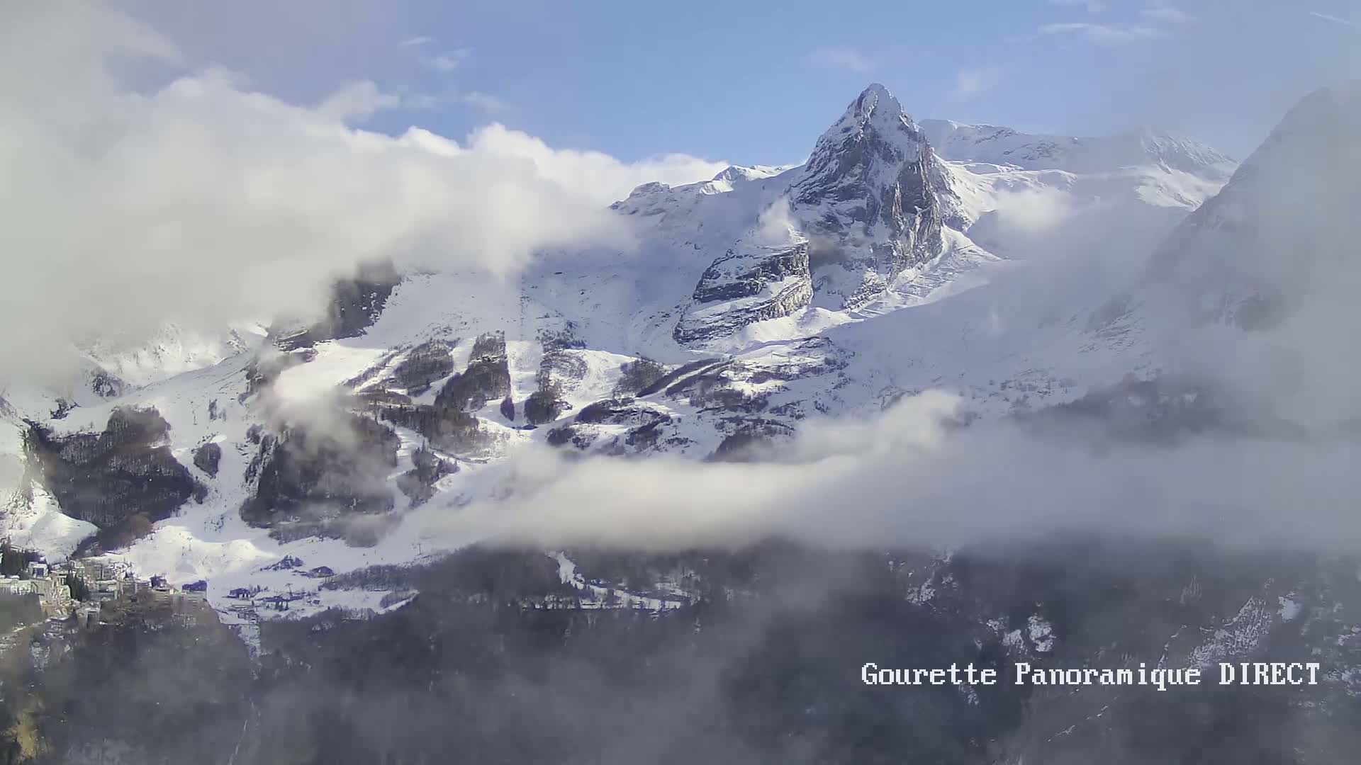 A snow-covered mountain range partially obscured by clouds under a mostly clear sky.