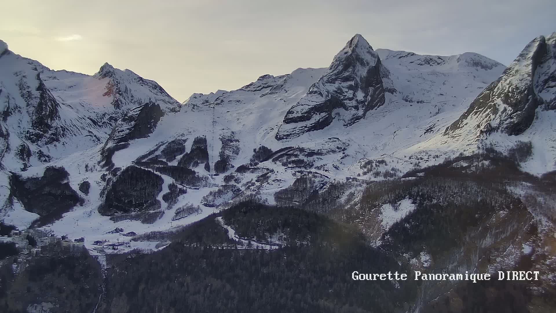 A snow-covered mountain range with a ski resort nestled in the valley below, under a partly cloudy sky.