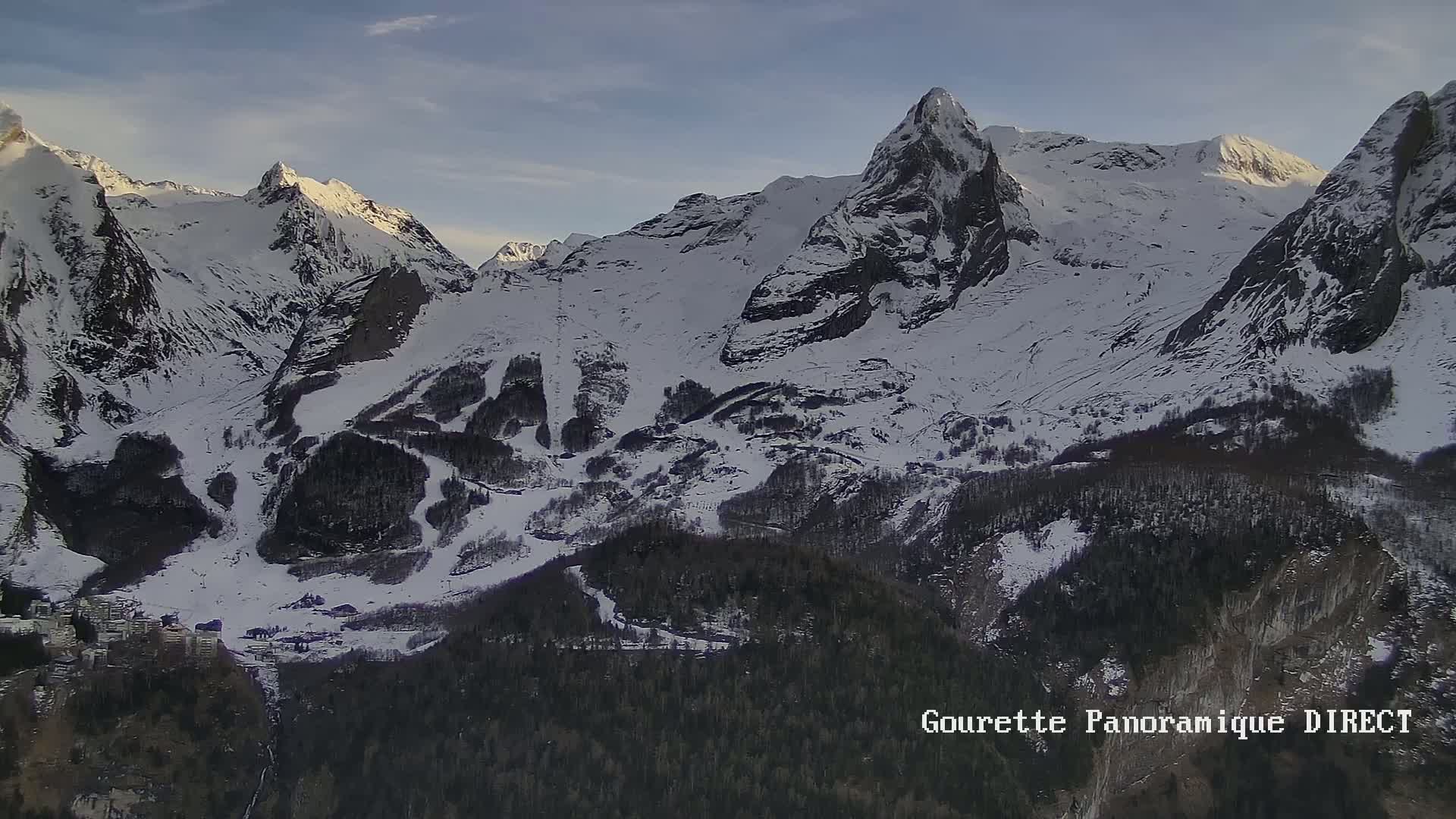 A snow-covered mountain range with dark evergreen trees at the base under a mostly clear sky.