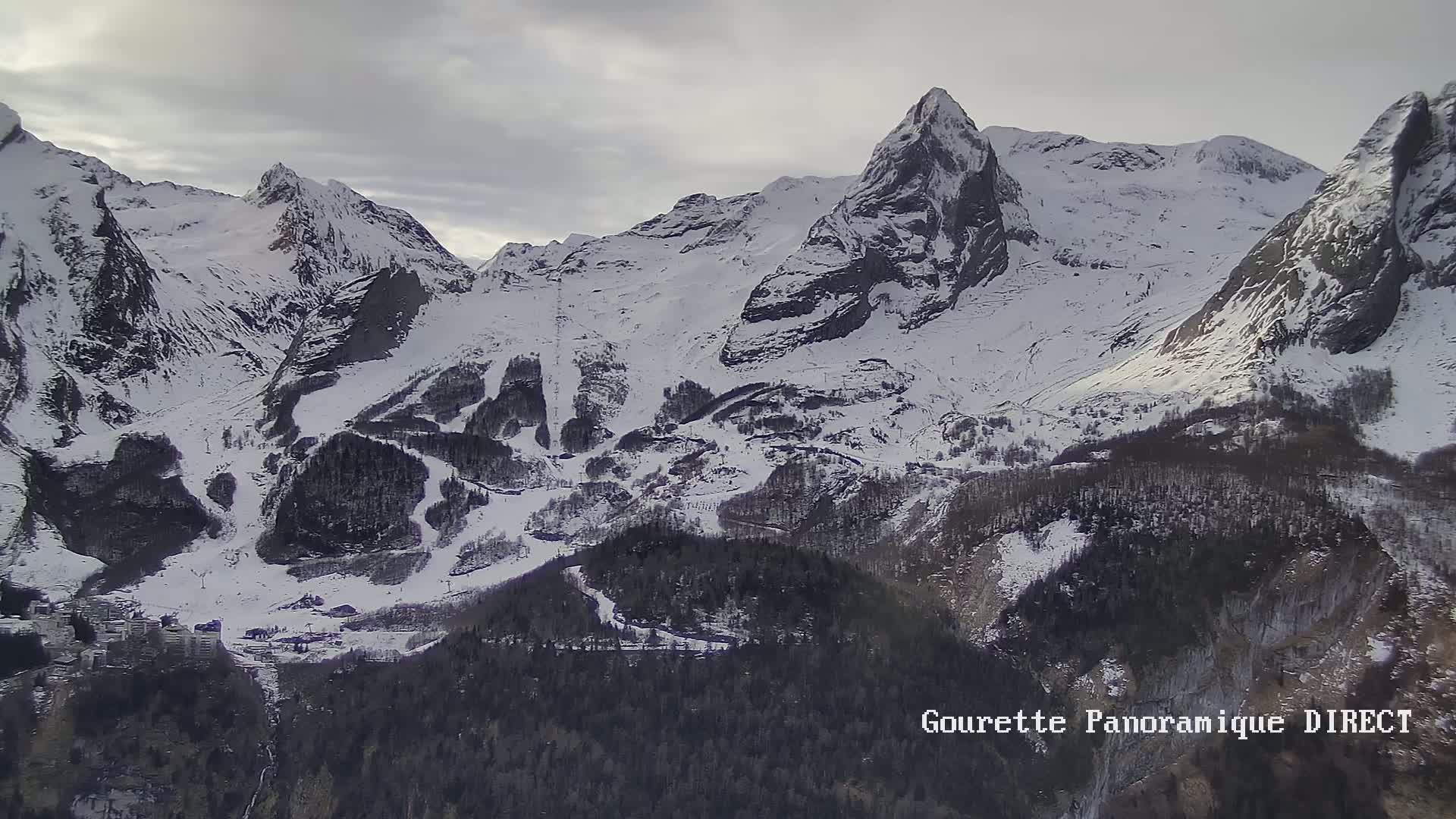 A snow-covered mountain range with dark evergreen trees below, under an overcast sky.