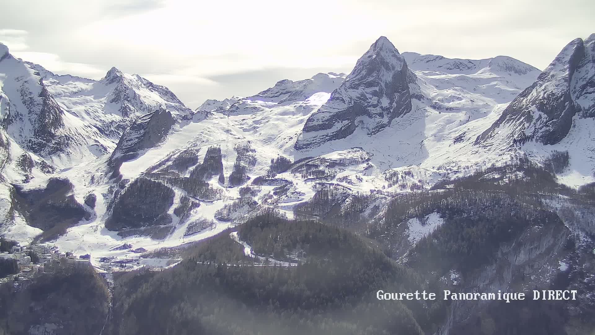 A snow-covered mountain range with ski slopes and a small village nestled in the valley below, under a mostly cloudy sky.