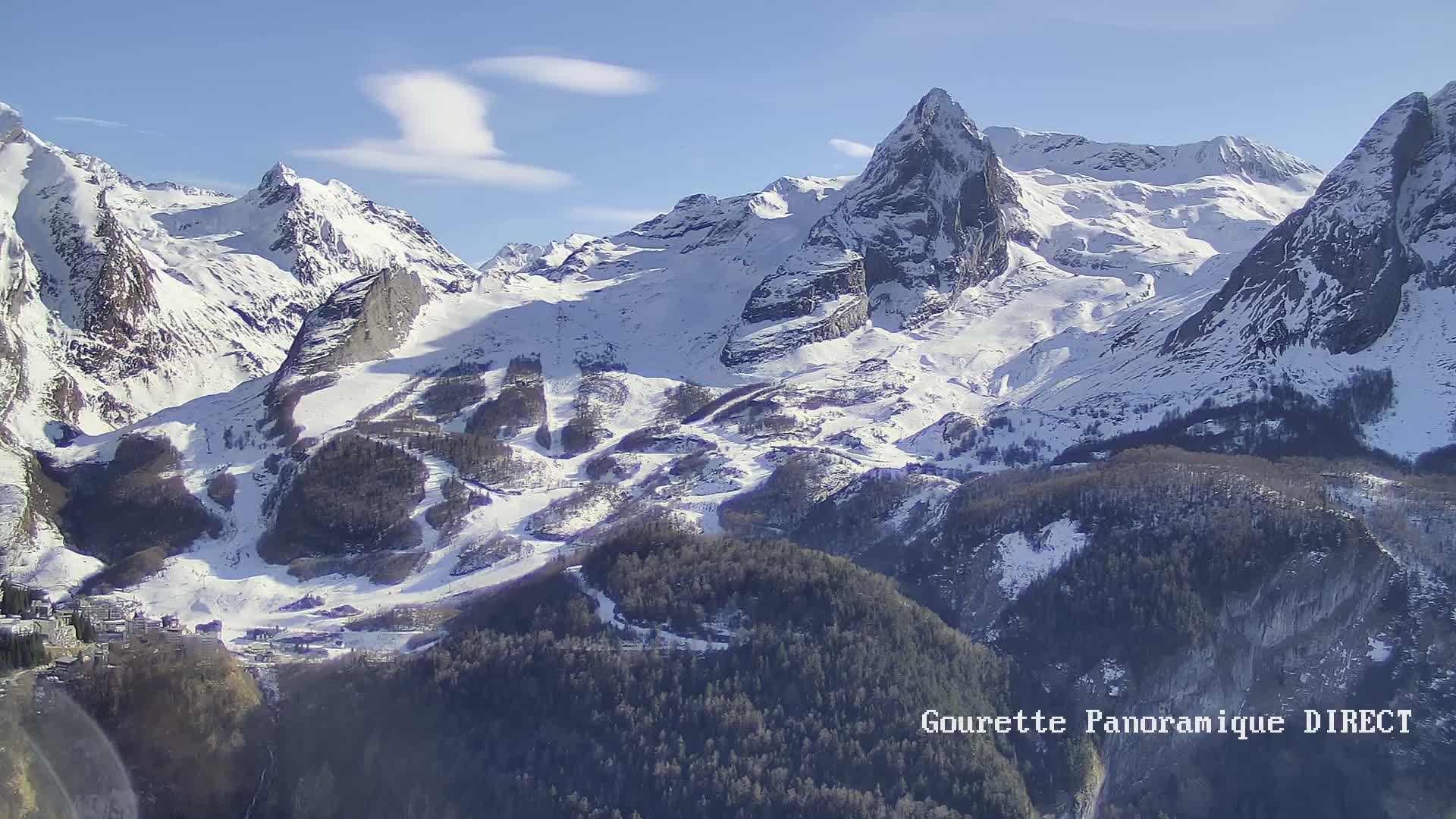 A snow-covered mountain range with a small town nestled in the valley below under a mostly clear blue sky.