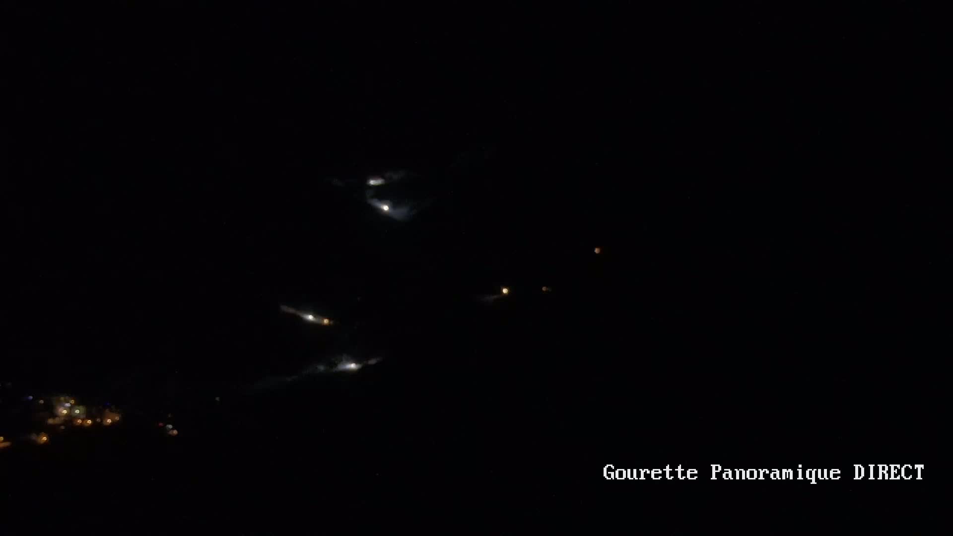 A nighttime long-range view shows scattered lights possibly from a town or city in the lower left corner and several brighter lights and streaks in the dark sky above.