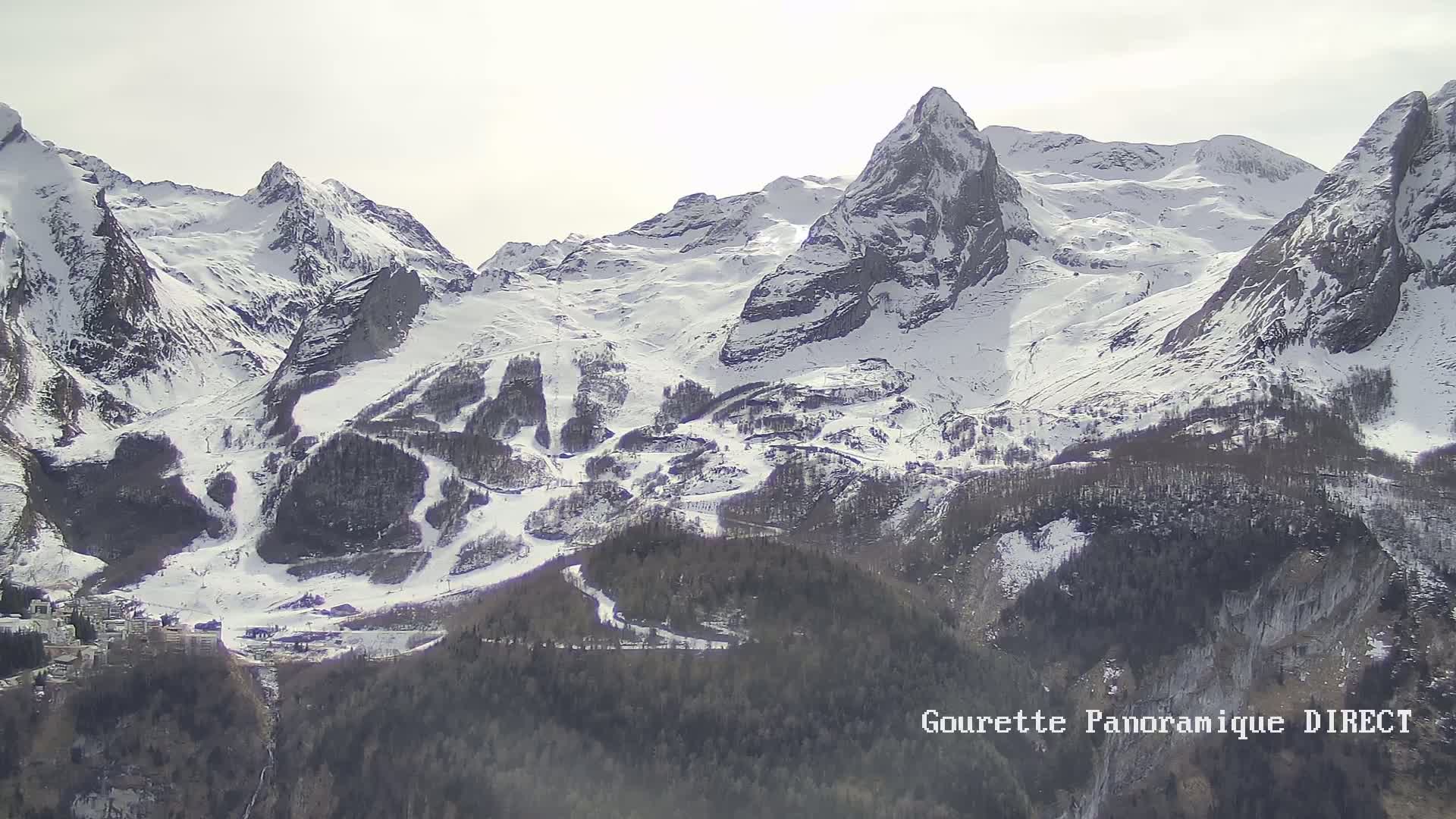 A snow-covered mountain range with ski slopes and a village nestled at its base under a bright, overcast sky.