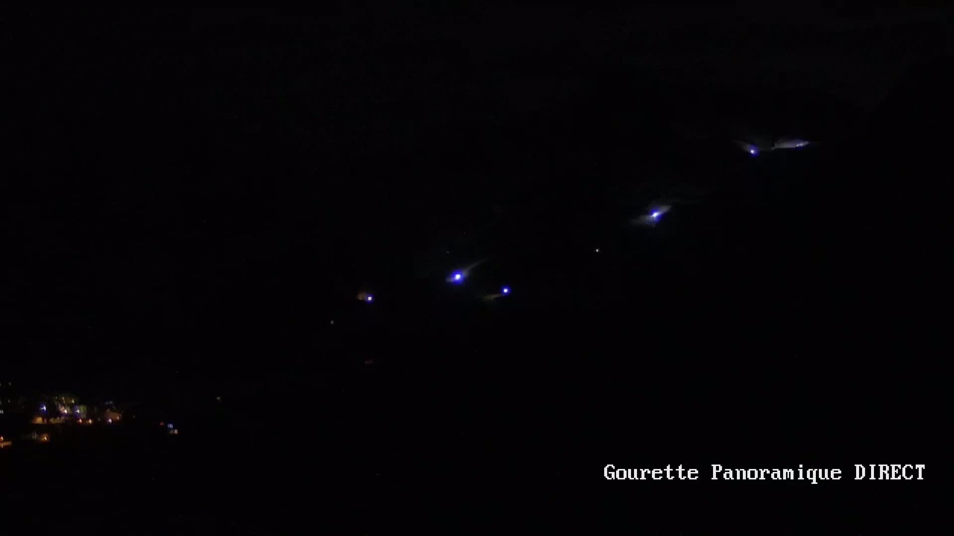 A nighttime, dark scene shows several faint blue lights in the sky above a dimly lit town.