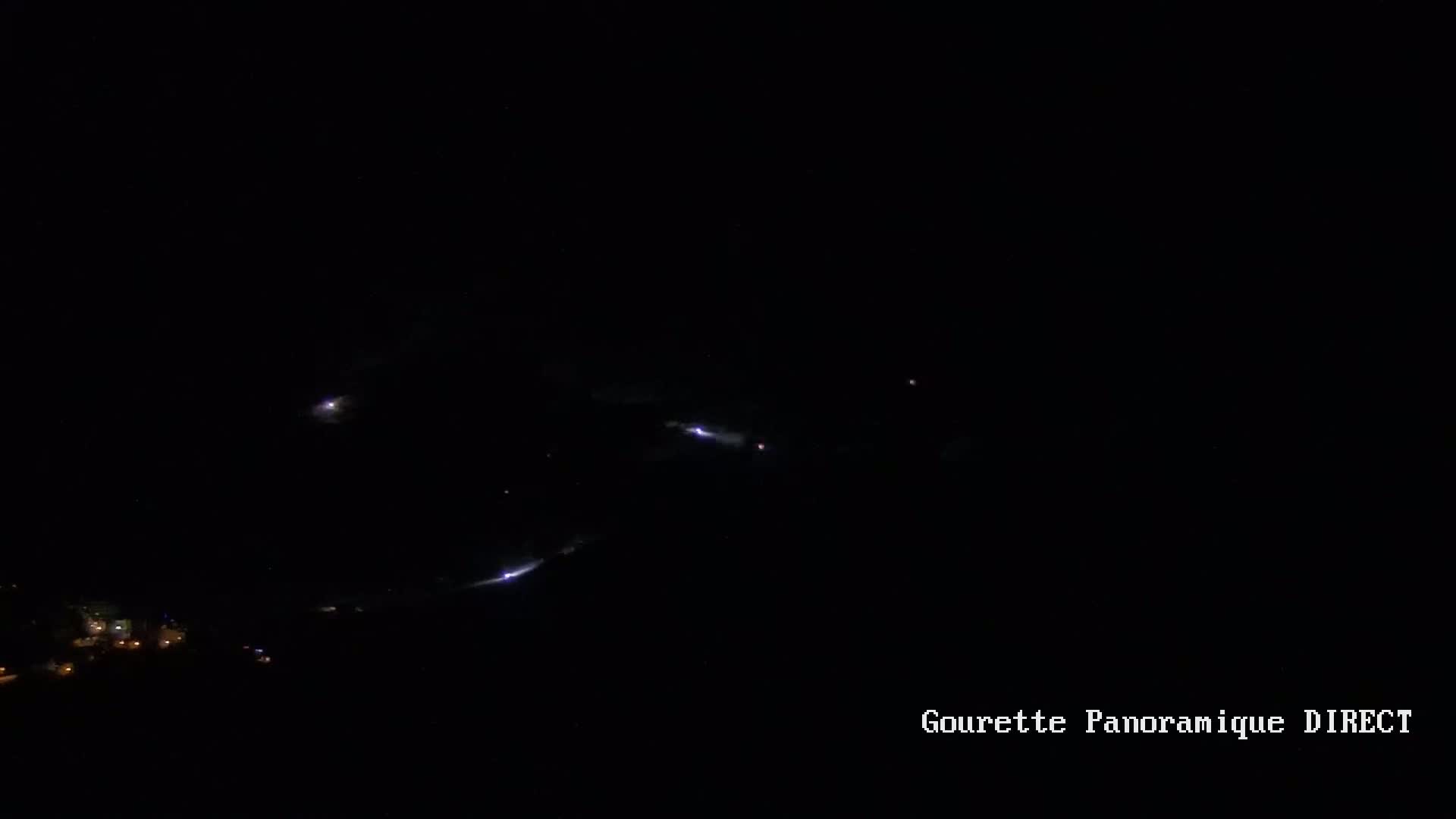 A nighttime, long-range view shows several streaks of lightning across a dark sky above a small cluster of lights on the ground.