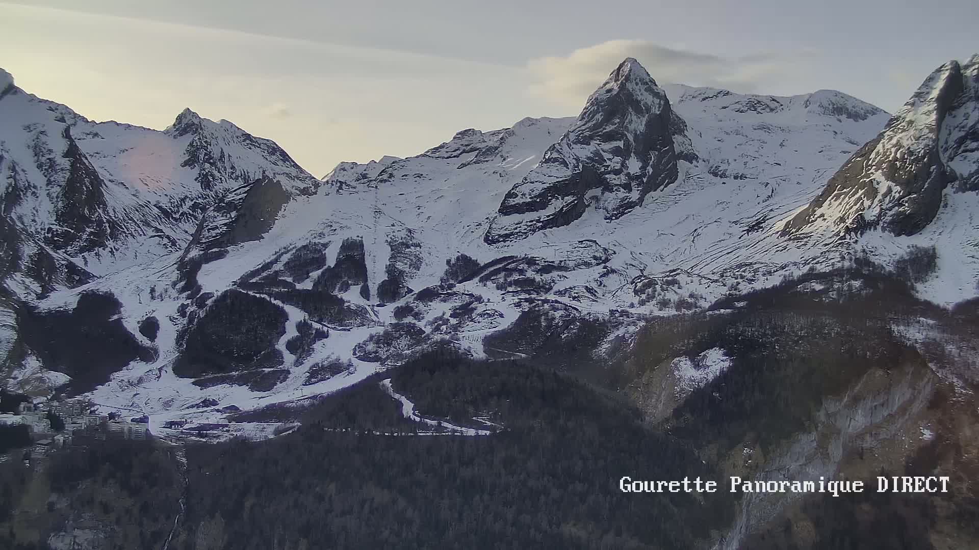 A snow-covered mountain range with a village nestled in the valley below under a partly cloudy sky.