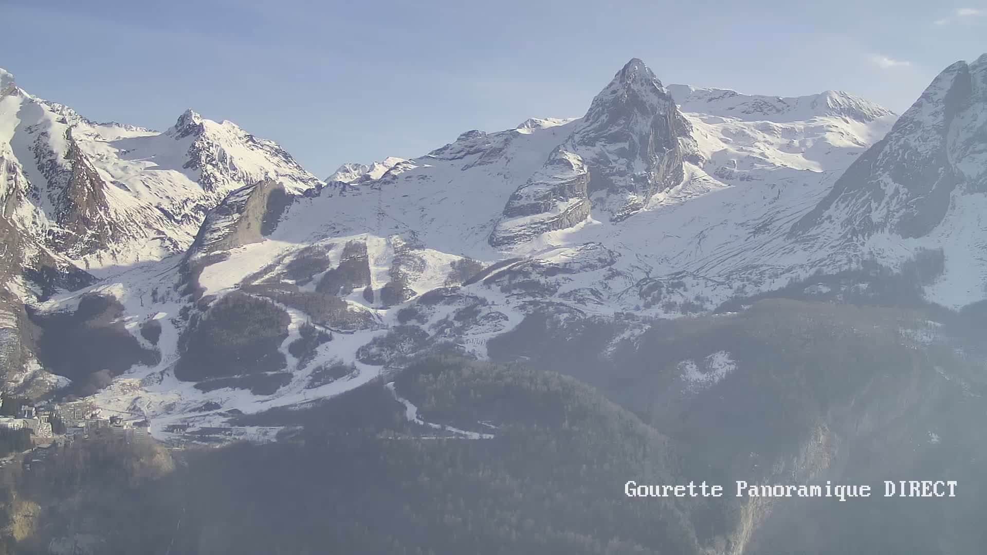A long-range view shows snow-covered mountains and a valley with a small town nestled below on a hazy, sunny day.