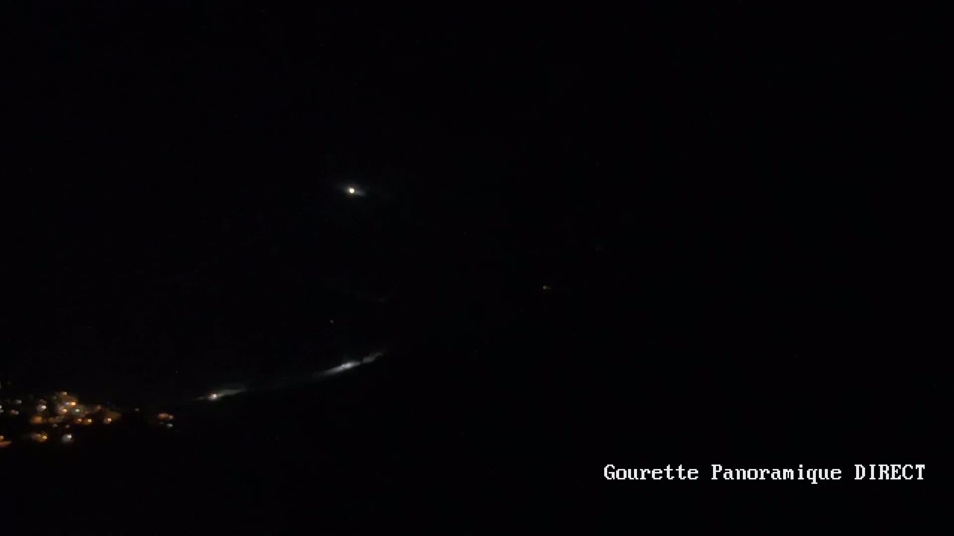 A nighttime long exposure shows a bright streak of light in the sky above a small town, with a bright moon visible.