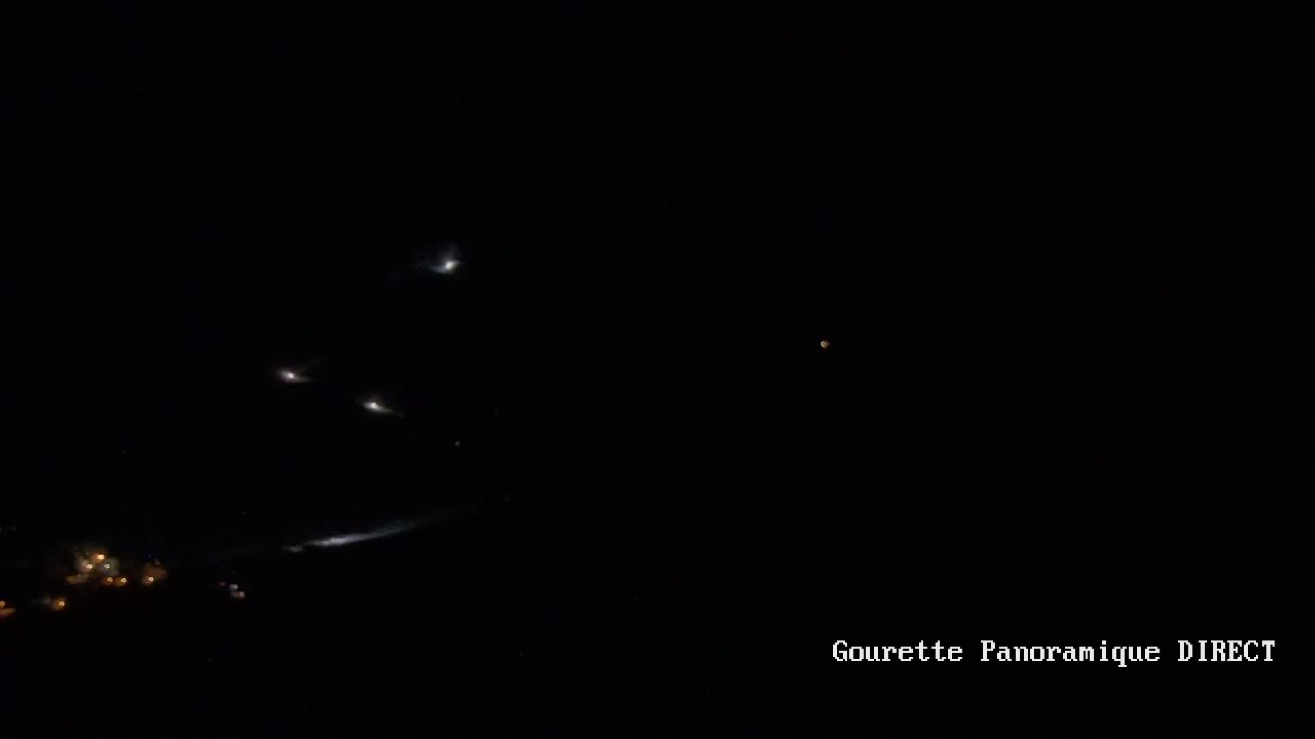 A nighttime long exposure shows bright lights streaking across a dark sky above a small cluster of lights on the ground.