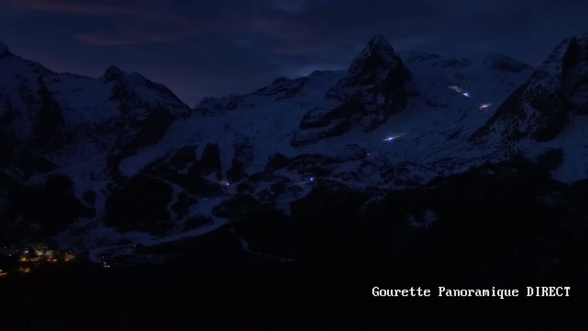 A nighttime, aerial view shows a snow-covered mountain range with a small village nestled at its base, illuminated by scattered lights.