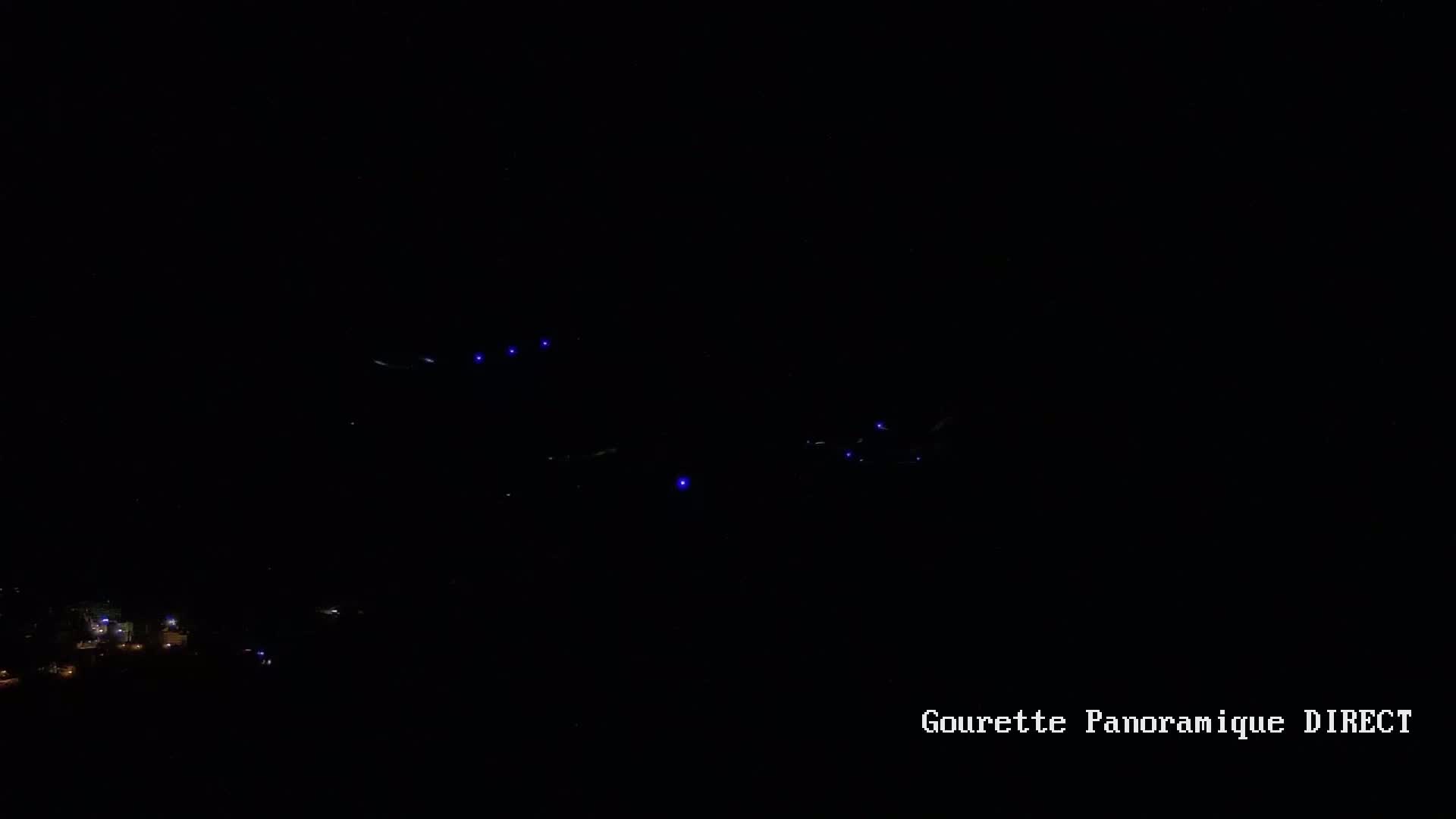 At night, several small, bright blue lights are visible in the dark sky above a dimly lit town.