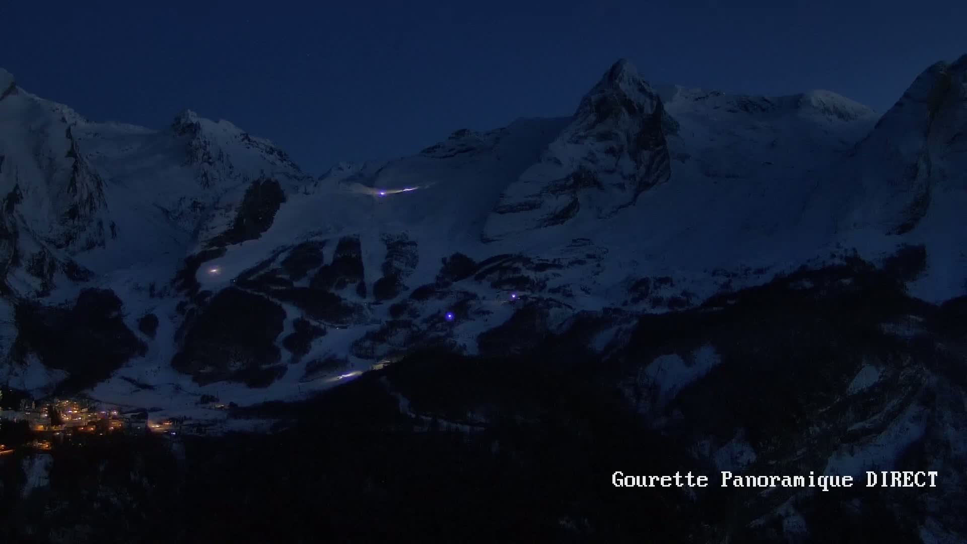 Snow-covered mountains at night, with a small village illuminated below and faint lights visible on the slopes.