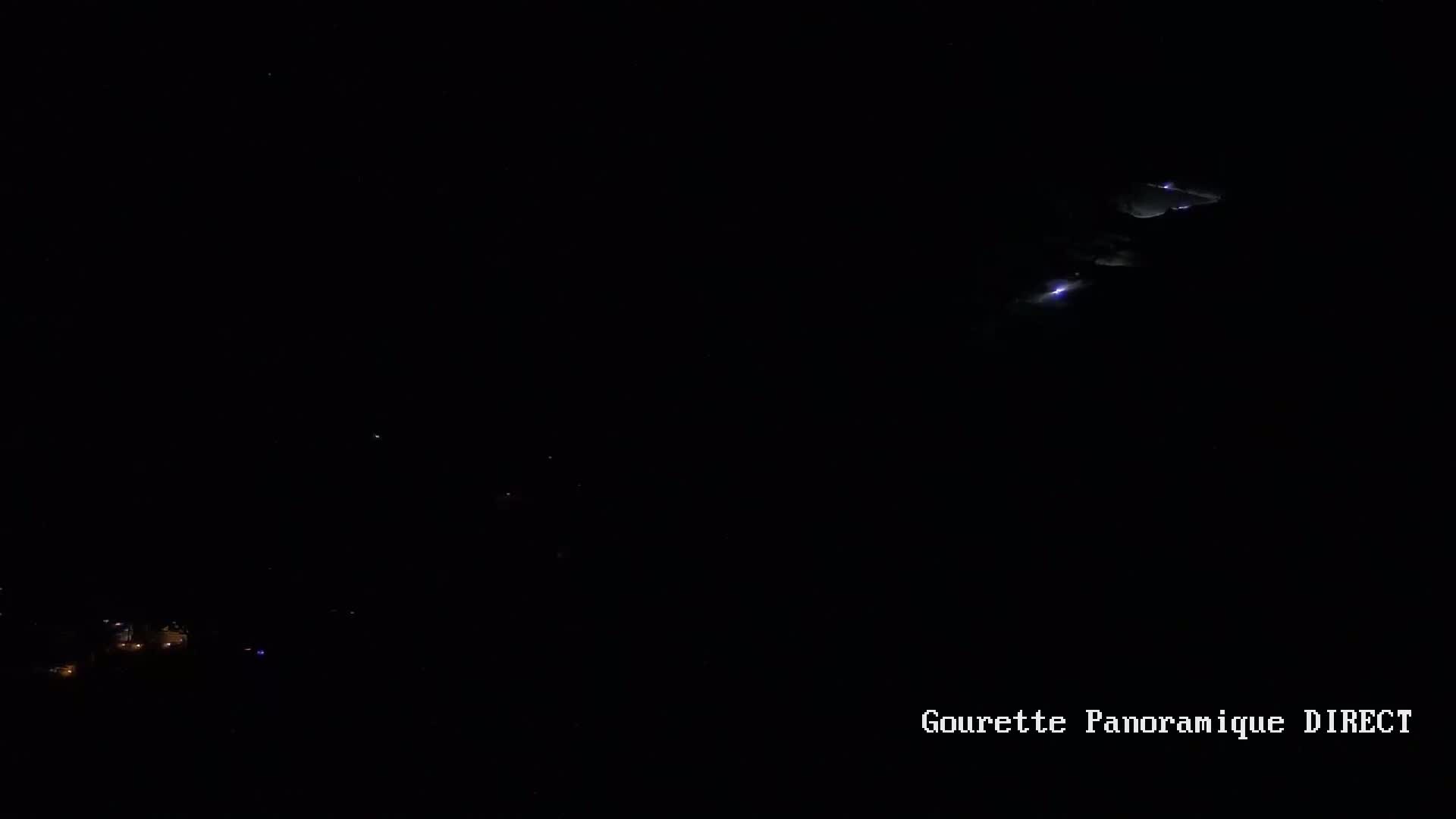 A nighttime, long-range view shows faint lightning strikes in the distance above a dark landscape with a few small lights visible near the bottom.