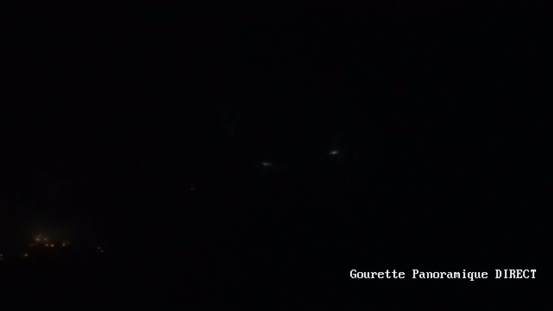 A nighttime long shot shows a dark sky with a few faint lights, possibly stars or distant lights on the ground, and a cluster of brighter lights in the lower left.