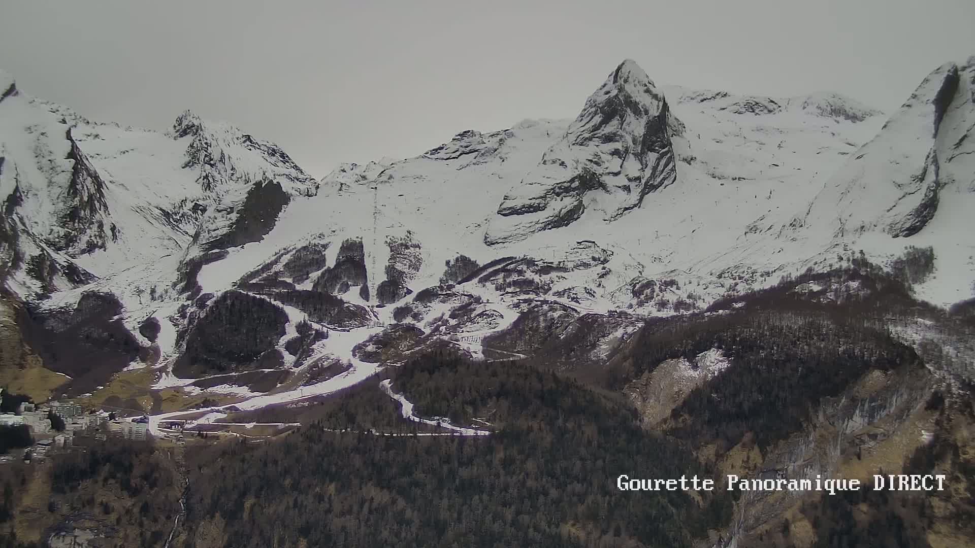 A snow-covered mountain range with ski slopes and a small town nestled at its base under an overcast sky.