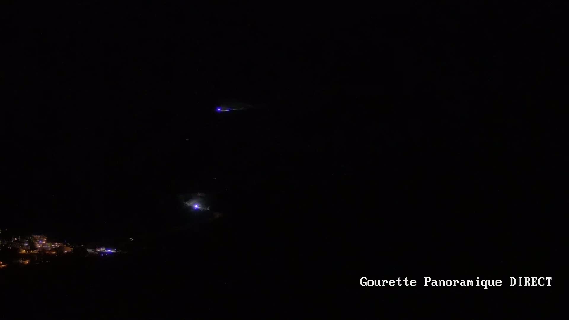 At night, faint purple lightning strikes illuminate a dark landscape with a small town visible in the distance.