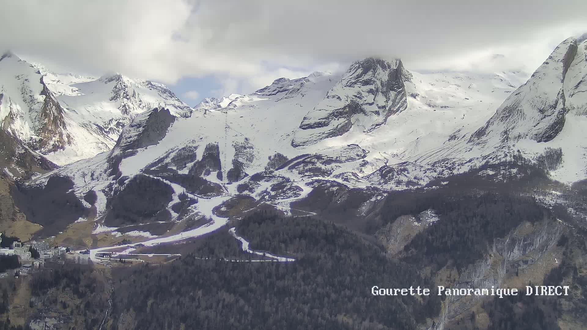 A snow-covered mountain range with ski runs visible, under a cloudy sky.