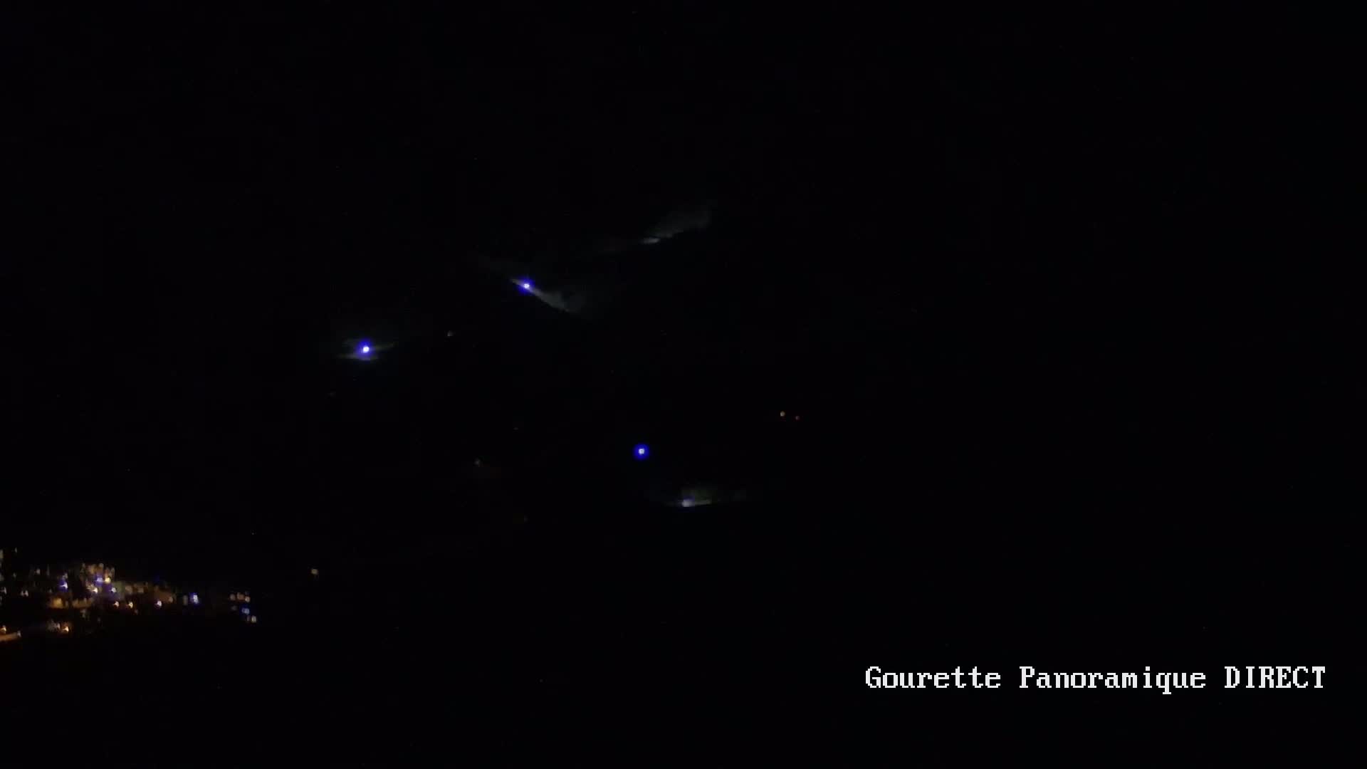 A nighttime long shot shows several faint blue lights in a dark sky above a small, dimly lit town.