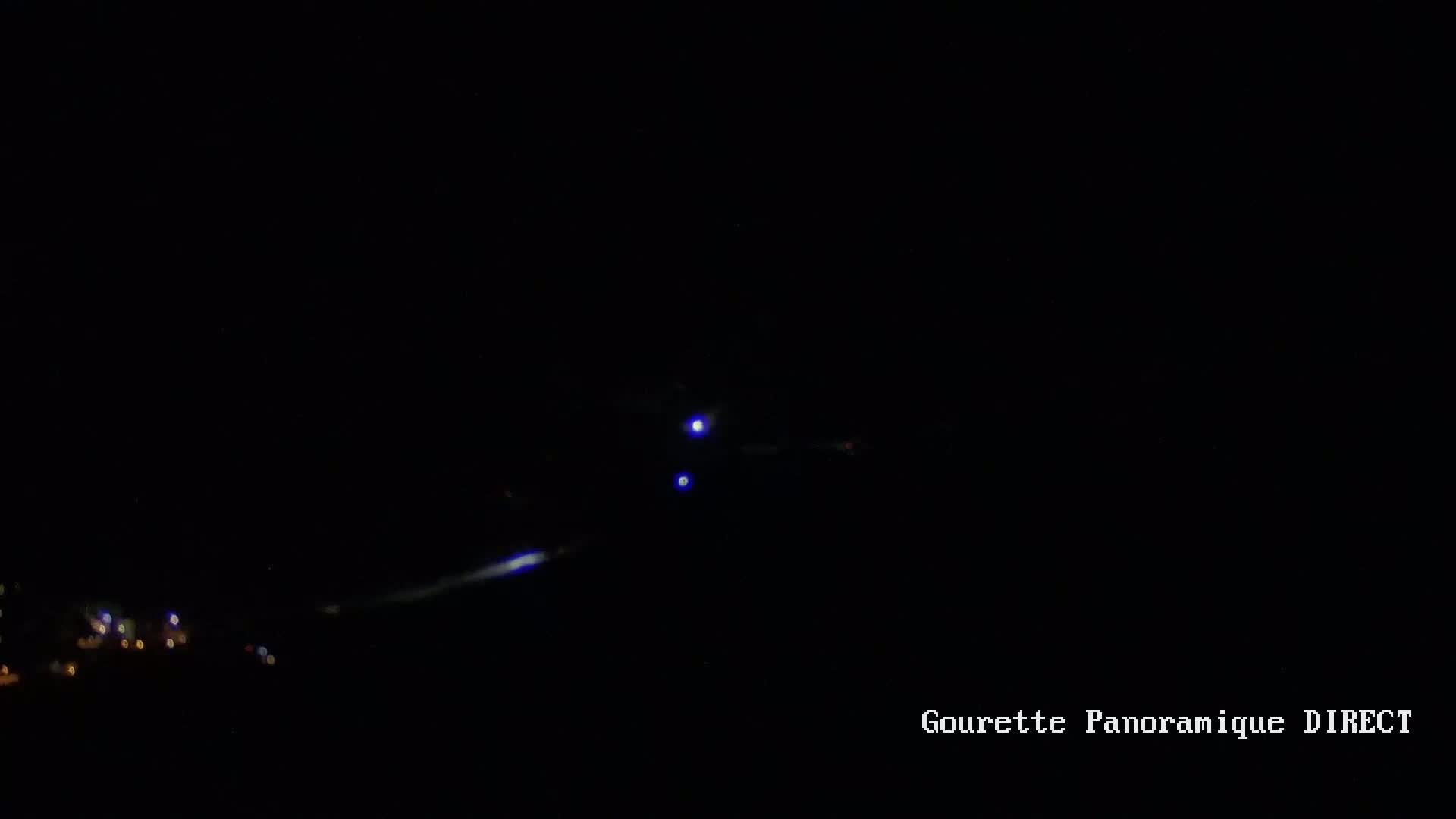 A dark nighttime scene shows two bright lights and a long streak of light in the sky above a cluster of dim lights on the ground.
