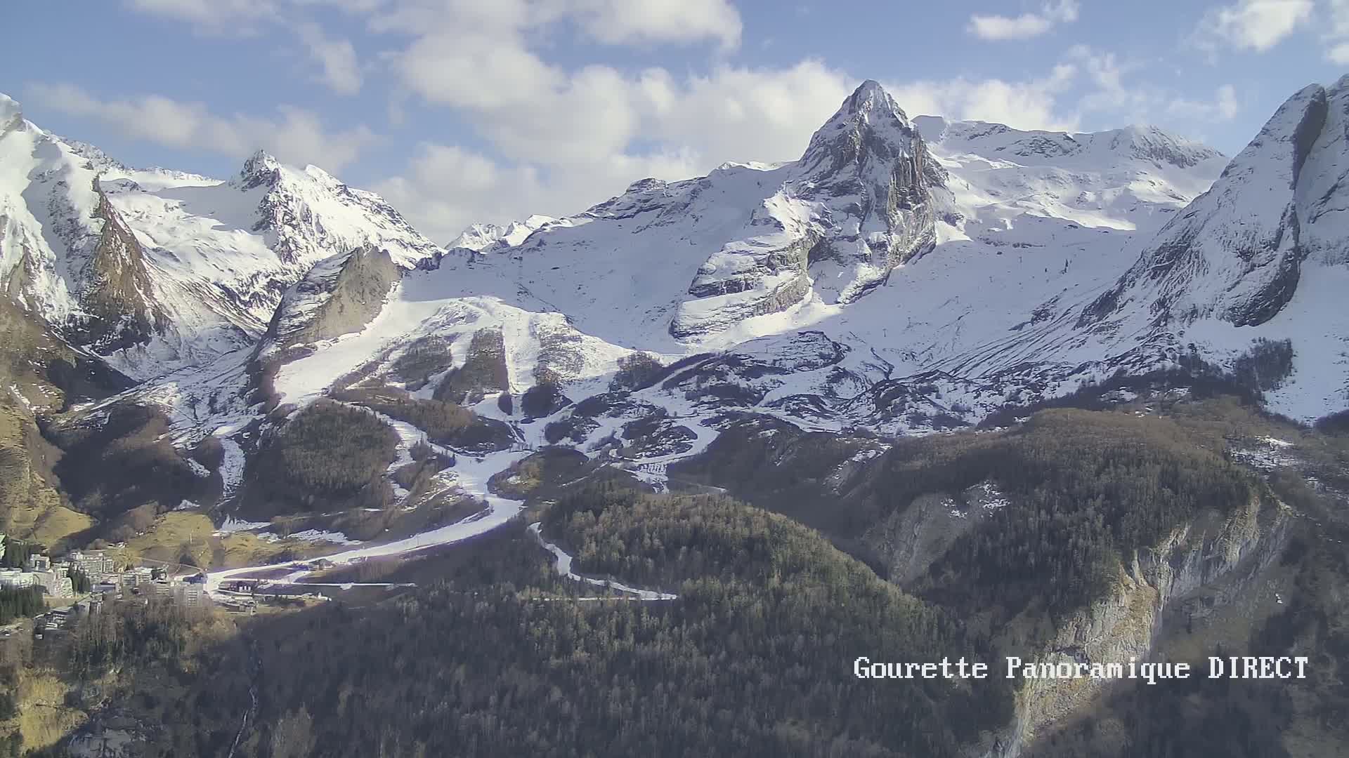 A snow-capped mountain range with ski slopes and a village at its base under a partly cloudy sky.