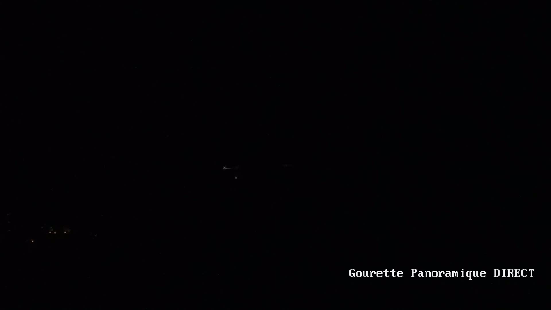 A dark nighttime scene shows a few faint lights in the distance and a very faint streak of light possibly from an airplane or other distant light source.