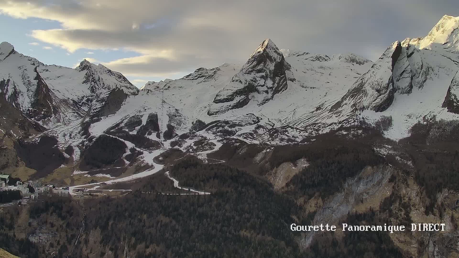 A long shot shows a snow-covered mountain range under a partly cloudy sky, with dark green forests below at the base of the mountains, and a small village nestled in the valley.