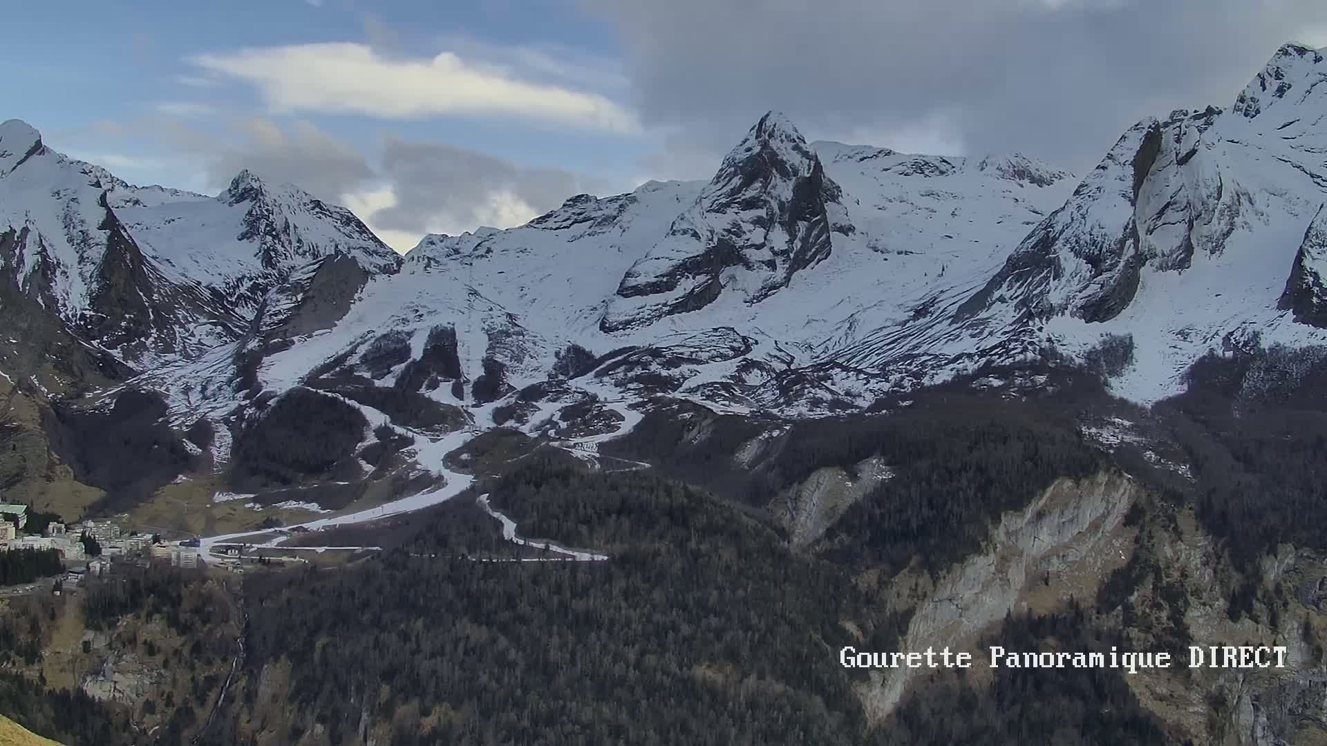 A snow-covered mountain range with a village nestled at its base under a partly cloudy sky.
