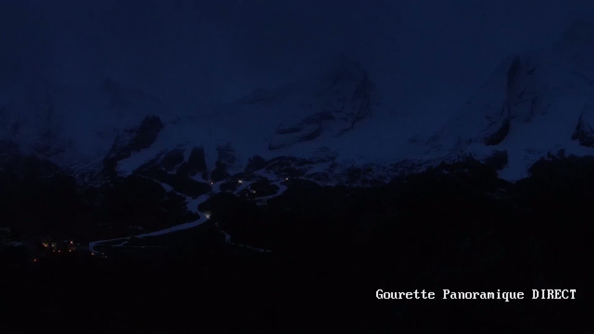 A dark, nighttime view of snow-capped mountains with a few faint lights illuminating a lower area.
