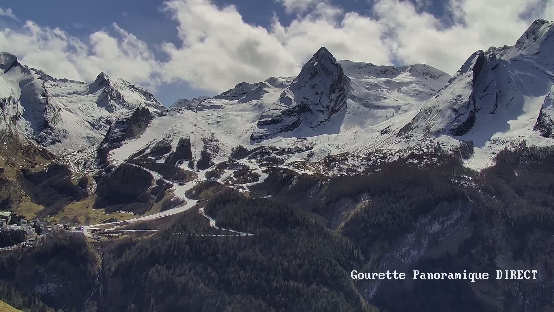 A long shot shows snow-covered mountains under a partly cloudy sky, with a village nestled in the valley below.