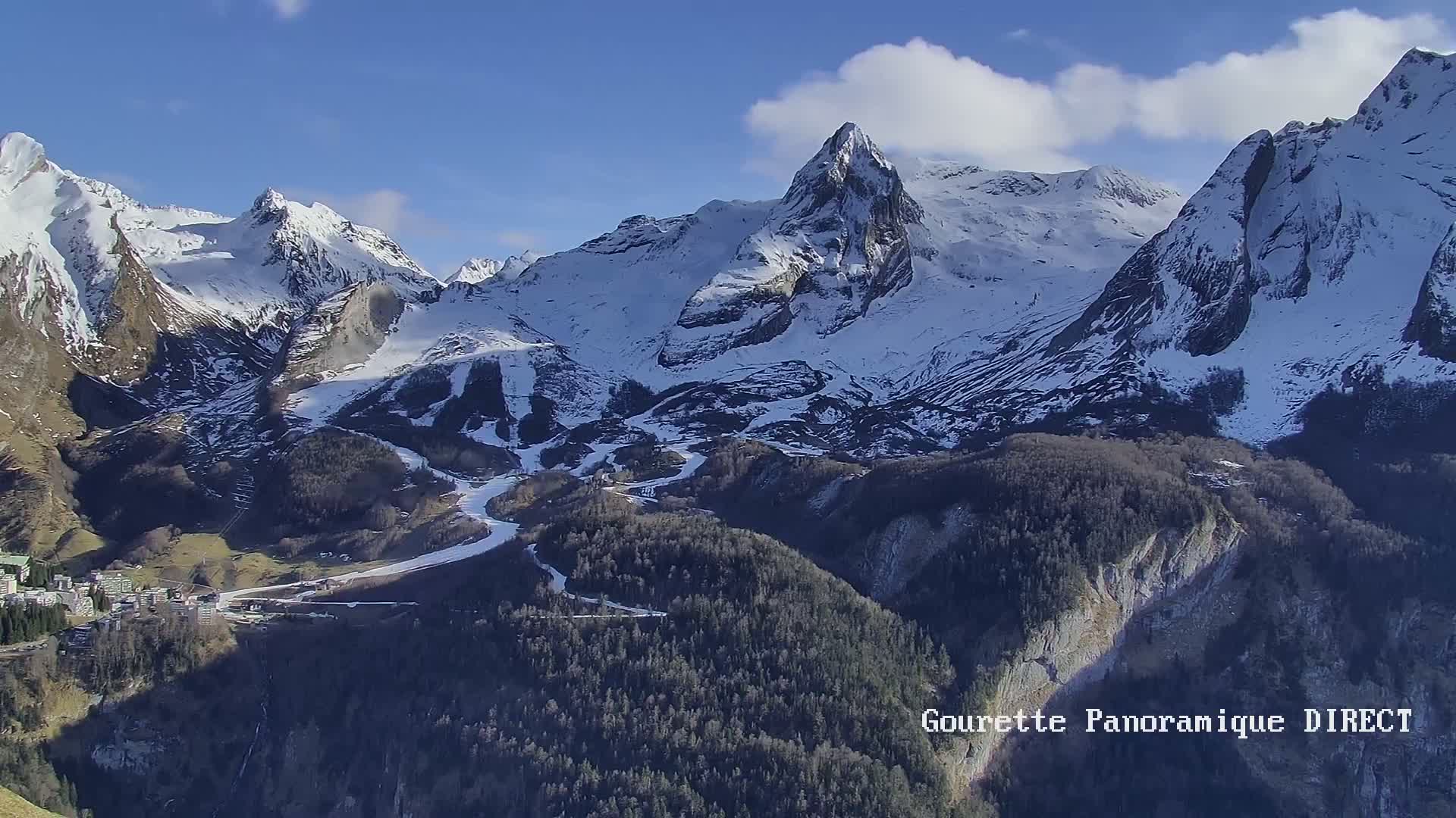 A snow-capped mountain range with a valley containing a small town and ski runs is visible under a mostly sunny sky.