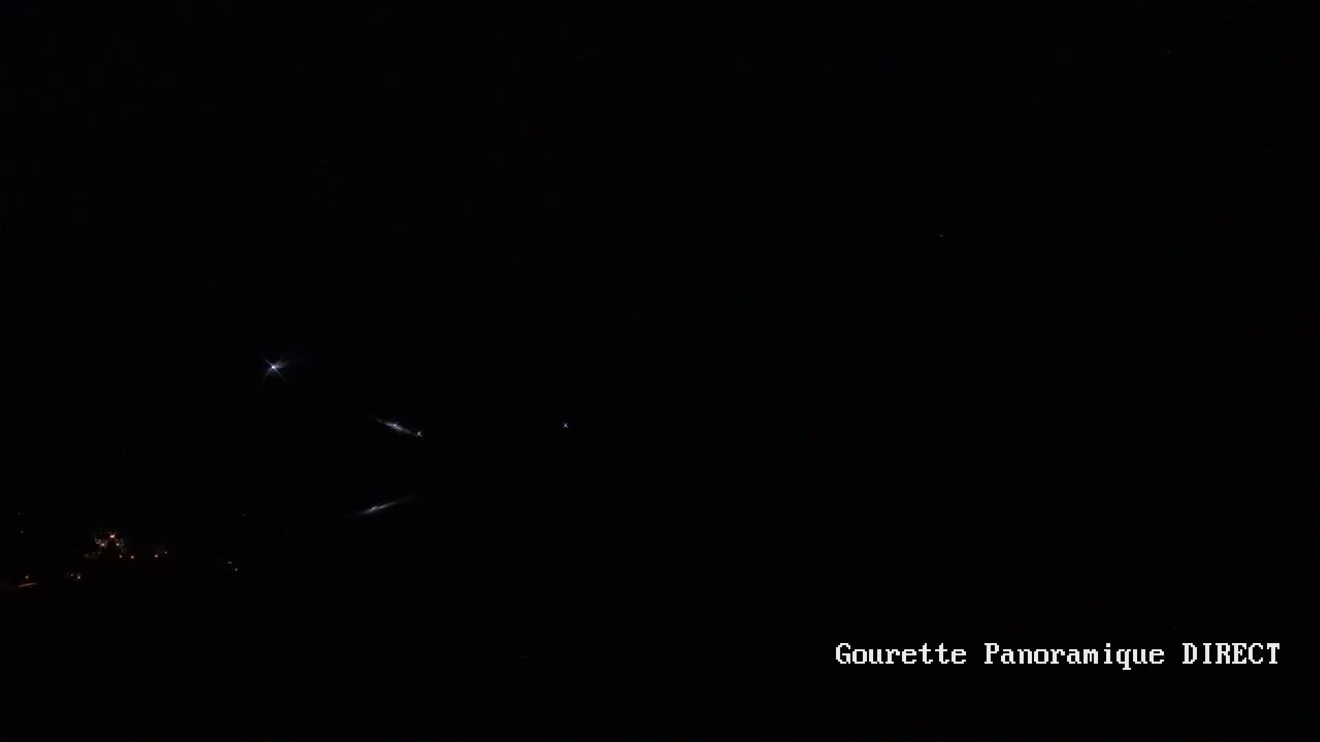 A nighttime long-exposure shot shows streaks of light in the dark sky above a small cluster of lights on the ground.