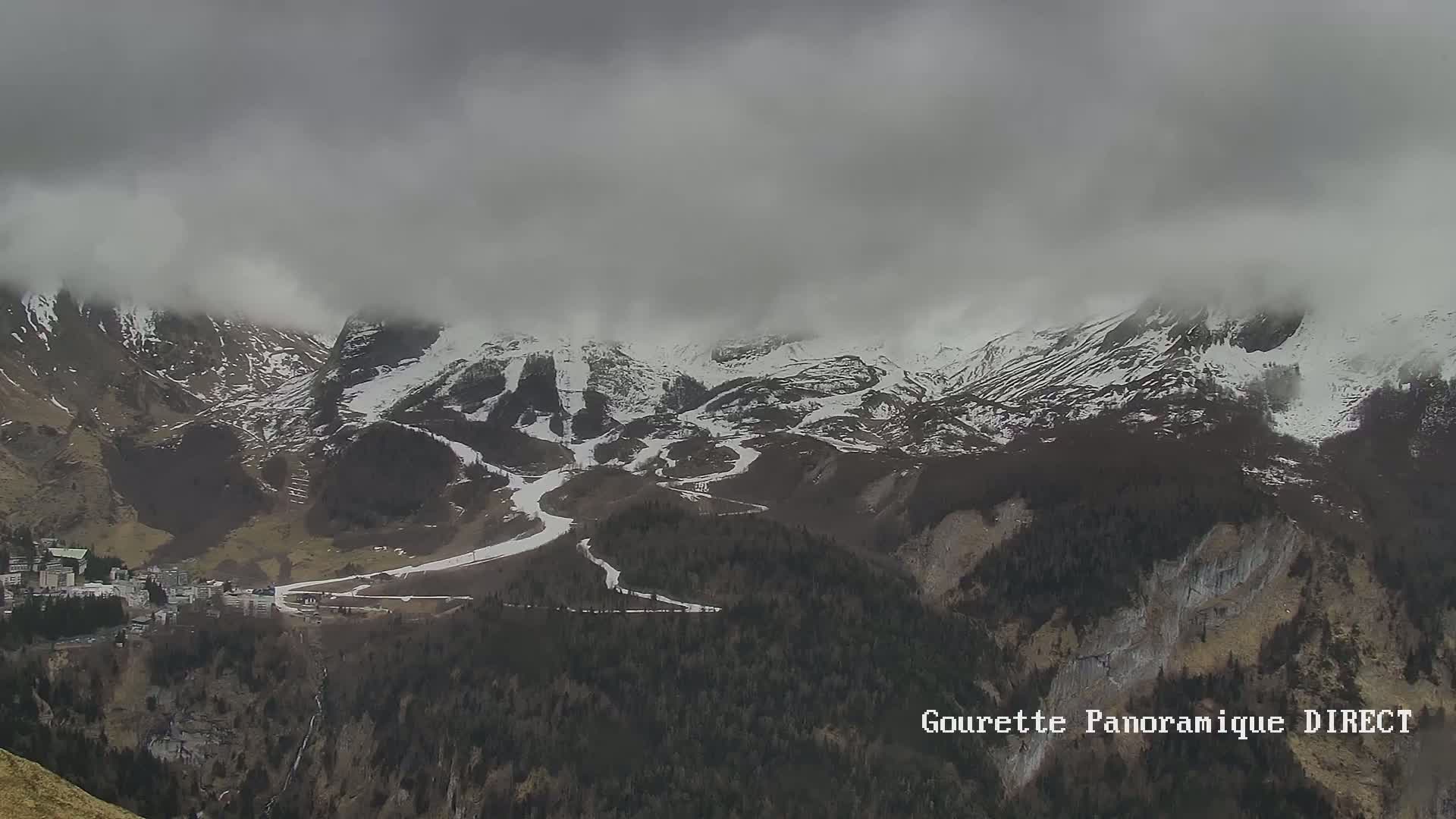 Snow-capped mountains partially obscured by a cloudy sky overlook a valley with a village and forested hillsides.