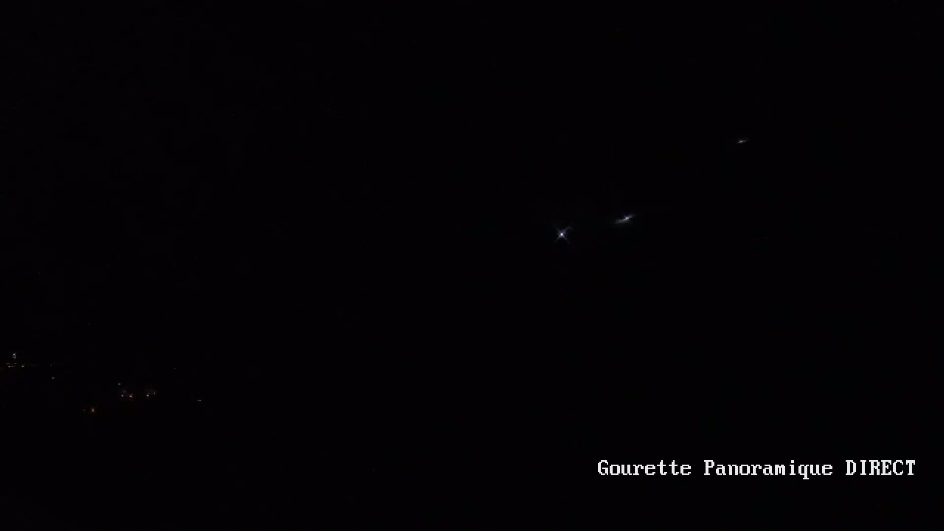 A dark nighttime scene shows a few bright lights in the distance, possibly airplanes, and faint lights below, possibly a town or city.