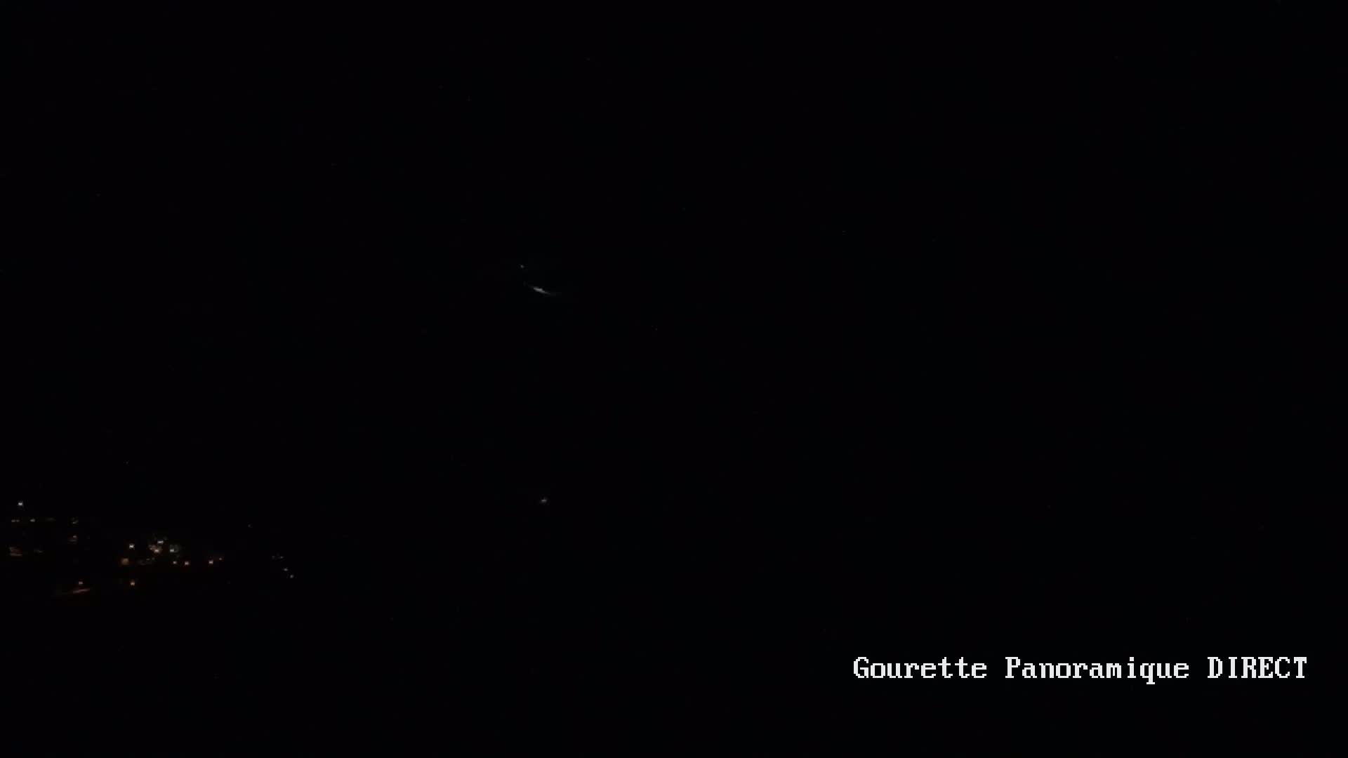 A nighttime long shot shows a faint, distant light source in a dark sky above a cluster of small lights on a dark ground.