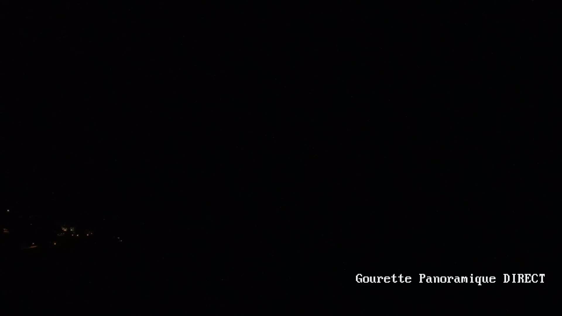 A dark nighttime scene shows a few distant lights on a hillside under a clear, black sky.