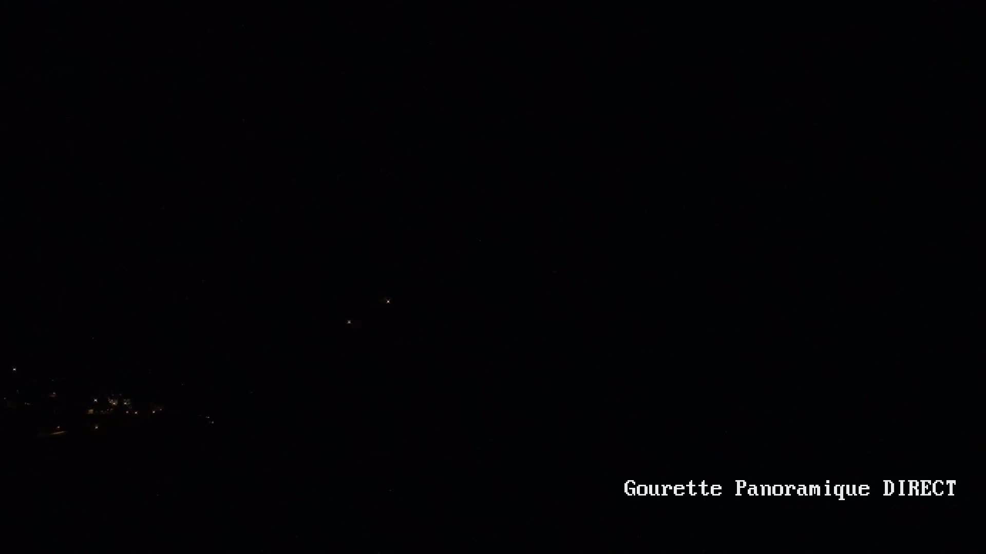 A nighttime view shows a few distant lights from a town or village below a dark, clear sky.