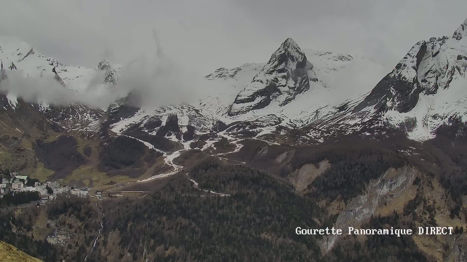 A long shot shows snow-capped mountains partially obscured by clouds, overlooking a valley with a small town and dark green forests.