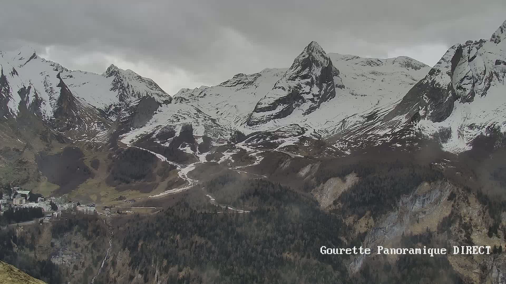 A long shot shows snow-capped mountains under an overcast sky, with a small town nestled in the valley below.